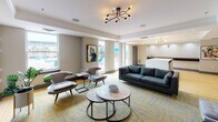 Located at 30 Maryland Ave in Rockville, Maryland, this single-floor lounge space combines contemporary design with inviting communal areas. The layout is thoughtfully organized into distinct zones, each serving a specific social function while maintaining a cohesive aesthetic throughout.

The main lounge area features plush, modern seating arrangements, including a deep charcoal sofa, accent chairs, and round marble-topped coffee tables set atop a vibrant blue and gold patterned area rug. A sputnik-style chandelier with exposed Edison bulbs provides a stylish focal point overhead, complemented by crown molding and recessed lighting.

A standout feature is the fireplace alcove, where a sleek gas fireplace is framed in light stone tile and topped with a wall-mounted flat-screen television. Bold geometric gold-and-white wallcovering accents the surrounding walls, adding a retro-modern flair. Curated artwork and live plants enhance the warmth of this nook.

Adjacent to the lounge is a bar-height counter with black metal stools and light wood flooring, suitable for casual gatherings or work. Large windows throughout flood the space with natural light and offer views of an outdoor pool area, connecting the interior to the exterior environment seamlessly.