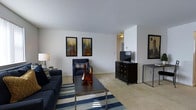 This single-floor space offers a thoughtfully designed interior that balances modern aesthetics with everyday comfort. The living area serves as the centerpiece of the space, featuring plush carpeted flooring that adds warmth underfoot. A navy blue sofa and matching accent chair create a cohesive seating arrangement, complemented by a sleek, metal-framed coffee table adorned with decorative copper and amber accents. A bold navy and white striped area rug anchors the seating area and adds a dynamic visual element to the room. The walls are finished in a clean, crisp white, providing a neutral backdrop that highlights a pair of framed cityscape art pieces, lending an urban sophistication to the space. A dark wood media dresser houses a flat-screen television, serving as a functional and stylish focal point along one wall. The open layout allows for a natural flow between areas, with a glimpse into an adjoining room visible in the background. Recessed and overhead lighting ensures the space is well-lit throughout. Overall, the interior presents a modern, coordinated aesthetic with a welcoming ambiance suited for both relaxing and entertaining.