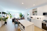 This well-designed 2-bedroom, 2-bathroom residence offers a clean, modern aesthetic throughout its single-floor layout. The open-plan kitchen and living area serves as the heart of the home, featuring a sleek white kitchen with flat-front cabinetry, open shelving, stainless steel appliances including a range, microwave, dishwasher, and refrigerator, and a central island with built-in lower shelving — ideal for both meal prep and casual dining. Light wood flooring flows seamlessly through the main living area, complementing the bright, white palette. The living room is generously proportioned, bathed in natural light from a bank of large windows that frame views of the surrounding neighborhood. The primary bedroom is a comfortable retreat, featuring a large window with outdoor views, a built-in closet, and soft carpeting. The second bedroom offers similar comfort and design continuity. Both bathrooms are finished with neutral tile, clean-lined white vanities, and large mirrors, with one featuring a tub-shower combination. Throughout the residence, recessed lighting and a crisp, contemporary design language create a bright and inviting atmosphere suitable for modern living.