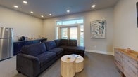 This well-appointed single-floor space offers an open, versatile layout designed for comfort and functionality. The main living area is anchored by a large sectional sofa in deep navy blue, complemented by natural wood cylindrical coffee tables that add a warm, organic touch. Recessed lighting illuminates the room evenly, while a set of French doors with transom windows floods the space with natural light and provides views to the outdoors. A wall-mounted flat-screen TV sits above a rustic wood media console, creating a clear entertainment zone at one end of the room. A grid of six framed art pieces depicting sky and abstract scenes adds a refined, curated feel to the neutral-toned walls. Adjacent to the living area, a compact dining setup features a light wood table surrounded by modern chairs, positioned beneath the artwork for a cohesive aesthetic. The space flows seamlessly through an open doorway into a dedicated kitchenette, which is equipped with dark cabinetry, granite-style countertops, a stainless steel full-size refrigerator, and counter space for small appliances. Soft carpet throughout ties the areas together, while clean white trim and light beige walls maintain a bright, inviting ambiance throughout the entire space.
