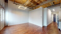 This single-floor loft space masterfully merges industrial character with contemporary design. The main open area immediately draws the eye upward to exposed wood-beam ceilings with visible black pipe infrastructure, lending an authentic warehouse aesthetic. Geometric recessed LED panel lighting provides a modern contrast against the warm, rustic ceiling overhead. Rich hardwood flooring runs throughout the primary living areas, adding depth and warmth to the expansive open layout.

A standout architectural feature is the original exposed brick wall, complete with a tall arched window framed in black that floods the space with natural light and serves as a striking focal point. The interplay between the raw brick and crisp white walls creates a compelling visual balance throughout.

Private rooms are finished with soft neutral carpet and access to the open corridor, offering flexibility for various uses. The kitchen is outfitted with white cabinetry, taupe countertops, a tiled backsplash, a matte black faucet, and stainless steel appliances, delivering a clean, functional workspace. The bathroom features large-format grey tile flooring, a walk-in shower with floor-to-ceiling tile surround, and black hardware accents that echo the industrial theme found throughout the space. Together, these elements create a cohesive, urban loft environment rich in character and detail.