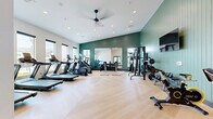 Located in Thornton, Colorado, this fitness center offers a thoughtfully designed workout space spread across a single open floor. The room is defined by striking sage-green vertical shiplap accent walls that contrast beautifully with crisp white walls and ceilings, creating a modern yet inviting atmosphere. Herringbone-patterned light wood flooring runs throughout, adding a refined aesthetic to the functional space.

The facility is comprehensively equipped to support a wide range of fitness activities. A row of treadmills lines one wall, accompanied by multiple elliptical trainers and a stair-climber for cardiovascular training. A dedicated area houses spin bikes alongside a stability ball, ideal for cycling and core workouts. The strength training section features a power rack, cable machine, dumbbells, and kettlebells, with a large wall-mounted mirror enhancing both functionality and the sense of space. A flat-screen TV is mounted on the accent wall for entertainment during workouts.

Natural light pours in through several large windows, offering views of the surrounding neighborhood and keeping the space bright and energizing. Recessed lighting and a ceiling fan provide additional comfort. The entrance features sleek glass-panel doors, and a water station is conveniently located along one wall. Overall, this fitness center combines practical amenities with polished design elements.