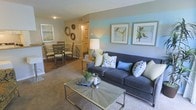 This single-floor residence offers a thoughtfully designed open-concept layout that connects the living, dining, and kitchen areas into one cohesive and inviting space. The living area is a standout feature, centered around a plush charcoal sofa adorned with an array of accent pillows in soft greens, blues, and neutrals. A geometric metal coffee table anchors the seating area, which rests on wall-to-wall carpeting that adds warmth underfoot. The accent wall behind the sofa is painted in a calming pale aqua hue, complemented by framed coastal bird artwork that reinforces the light, breezy aesthetic throughout. Natural and ambient lighting work together to keep the space feeling bright and welcoming. The dining area sits adjacent to the living room and features a round glass-top table surrounded by cushioned chairs, creating an intimate setting for meals. Circular wall-mounted decorative mirrors add visual interest to the dining wall. Beyond the dining space, a glimpse into the kitchen reveals warm cabinetry and additional lighting, suggesting a functional and well-appointed cooking area. The overall ambiance is relaxed yet polished, with a consistent coastal color palette of blues, greens, and warm neutrals tying each area of the home together harmoniously.