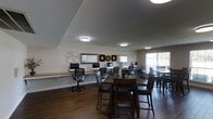 This single-floor community space is thoughtfully designed to serve multiple functions, combining a computer lab and social gathering area in one cohesive room. Along one wall, a long wooden workstation desk hosts a row of desktop computers accompanied by ergonomic mesh office chairs, providing residents with convenient access to technology. Above the workstation, decorative wall accents — including ornamental wreaths and circular metal artwork — add a touch of personality and warmth to the space.

The central and far areas of the room are furnished with dark wood high-top dining tables and matching chairs, offering comfortable seating for meals, meetings, or casual conversation. Natural light filters in through large windows fitted with horizontal blinds along the right wall, brightening the room alongside multiple flush-mount ceiling light fixtures. The ceiling features a distinctive tray design accented with crown molding, lending an element of architectural character to the space.

Rich, dark hardwood flooring runs throughout, providing a warm contrast to the light-toned walls. The textured ceiling and neutral color palette create a relaxed, inviting ambiance, making this a versatile and functional community hub suited for both individual use and group activities.