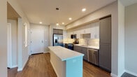 This single-floor unit showcases a contemporary design with clean lines and a neutral color palette throughout. The kitchen is a standout feature, offering a galley-style layout with two-tone cabinetry — white upper cabinets paired with gray lower cabinets — complemented by a tile backsplash and bright white quartz countertops. A central island provides additional prep space and casual seating, while a full suite of stainless steel appliances, including a French door refrigerator with ice maker, gas range, over-the-range microwave, and dishwasher, equips the space for modern living. Rich, dark hardwood-style flooring runs throughout the kitchen and living areas, adding warmth and depth to the space. The bedrooms are well-proportioned, each featuring ceiling fans, soft neutral wall tones, and windows fitted with horizontal blinds that allow for natural light control. One bedroom offers views of the surrounding landscape. The bathroom is finished with a gray vanity, white quartz countertop, a large framed mirror with vanity lighting, and a full bathtub with shower. Recessed lighting throughout ensures a bright and welcoming ambiance across all areas of the unit.