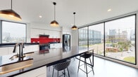 This contemporary single-floor space is defined by a thoughtfully designed open-plan kitchen and dining area that combines bold aesthetics with functional efficiency. The kitchen is anchored by an extensive run of flat-front white cabinetry, providing generous storage along the full length of the wall. A striking red subway tile backsplash serves as the focal point, adding a vibrant contrast against the crisp white surfaces and dark stone countertops. Stainless steel appliances, including a French door refrigerator and a range with oven, are seamlessly integrated into the layout. A large central island with a dark countertop and built-in bar seating doubles as a prep and dining surface, illuminated by a pair of stylish dome pendant lights overhead. Polished concrete floors run throughout the space, lending an industrial-modern character. Floor-to-ceiling windows and a full-height glass curtain wall flood the interior with abundant natural light and frame sweeping views of the surrounding urban skyline and streetscape. A secondary window positioned along the kitchen wall further enhances the brightness and connection to the outdoors. The space also features a glass-paneled double door leading to an adjacent room, adding both functionality and visual continuity to the overall layout.