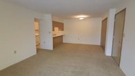 This single-floor unit features an open-concept layout that connects the living area to the kitchen through a functional pass-through design. The spacious living area is finished with wall-to-wall neutral beige carpeting, offering a clean and understated aesthetic. Walls are painted in a crisp white throughout, contributing to a bright and airy feel, complemented by a ceiling-mounted light fixture that provides ambient illumination.

The kitchen is partially separated from the living space by a raised countertop bar with wood-toned laminate cabinetry below, creating a natural division while maintaining visual openness. The kitchen includes white appliances and additional cabinetry along the walls, providing functional storage and workspace. A separate hallway leads further into the unit, where a door — likely to a closet or additional room — is visible along the far wall.

The layout is straightforward and practical, with clear sightlines across the main living space. The neutral color palette and simple finishes create a versatile environment easily adaptable to various furnishing styles. Overall, the space offers a functional and comfortable single-level living arrangement.