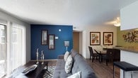 This single-floor residence offers an open-concept layout that seamlessly connects the living and dining areas, creating a cohesive and functional flow throughout the space. The living area is anchored by a striking deep blue accent wall, which serves as a bold focal point and adds a sense of depth and character to the room. Decorative framed artwork adorns the walls, complementing the contemporary aesthetic. A spacious sectional sofa with patterned throw pillows faces a dark wood coffee table, providing a comfortable gathering area atop a geometric area rug.

Wood-style plank flooring extends throughout the main living space, offering a warm, durable foundation that ties the rooms together. The textured ceiling adds a classic architectural touch overhead, while a visible HVAC vent ensures year-round climate control. The dining area, positioned adjacent to the living room, accommodates a rectangular table with seating for multiple guests, accented by additional framed artwork. A hallway leading to additional areas of the home is visible in the background, hinting at further living spaces beyond the main open area.