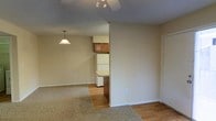 This single-floor residential space offers a practical and comfortable layout designed to maximize functionality. The open living area is finished with warm, neutral-toned walls that create a welcoming ambiance. Underfoot, a combination of plush carpeting and hardwood-style flooring defines the distinct zones of the space, with carpet anchoring the living area and hard flooring transitioning seamlessly into the kitchen.

The kitchen is neatly tucked adjacent to the main living area and features warm-toned wooden cabinetry, a white refrigerator, and a countertop with a granite-style finish. A pendant light fixture hangs above, adding a touch of character to the cooking and dining zone.

The living area benefits from ceiling-mounted track lighting as well as a ceiling fan, providing both focused illumination and climate comfort. Clean white baseboards and door trim provide a crisp contrast against the neutral walls, contributing to a tidy, well-maintained aesthetic throughout.

Overall, the space presents a straightforward and functional interior, suited for everyday living, with cohesive design elements that lend it a modest yet comfortable character.