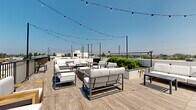 Located at 4971 S Centinela Ave in the Mar Vista neighborhood of Los Angeles, this rooftop deck offers an expansive open-air experience designed for both relaxation and social gatherings. The deck is finished with wood-look porcelain tile flooring throughout, lending a warm, contemporary aesthetic that complements the clean white parapet walls and sleek black metal railings that frame the perimeter.

At the heart of the rooftop is a generously sized lounge area furnished with modern outdoor sofas and chairs in white cushioning with dark metal frames, centered around a built-in rectangular fire pit — an ideal setting for evening gatherings under the open sky. String lights are strung across the entire space, creating an inviting ambiance after dark.

Raised concrete planters filled with drought-tolerant succulents and lush greenery add a natural, living element to the rooftop's design. A dedicated walkway with tiled flooring runs along the perimeter, providing unobstructed panoramic views of the surrounding Los Angeles cityscape, palm trees, and open sky in every direction. The elevated vantage point captures the breadth of the neighborhood and beyond. The overall design strikes a balance between functional outdoor amenity space and a stylish, resort-like atmosphere suited to the Southern California lifestyle.