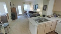 This single-story space showcases an open-concept design that seamlessly connects the kitchen and living area, creating a cohesive and functional layout. The kitchen features a peninsula with a double-basin stainless steel sink and light-toned cabinetry, providing ample storage and counter space while maintaining a clear sightline into the living area. Neutral countertops complement the warm, understated color palette found throughout the space. The living area is carpeted in a plush, light-toned textile that adds warmth underfoot. Large windows and a glass-paneled door flood the space with natural light, offering views of the outdoors and enhancing the open, airy ambiance. Sheer green-accented curtains frame the windows, adding a subtle touch of color without disrupting the room's soft, neutral tone. Framed artwork on the walls contributes to the finished, well-appointed feel of the space. The overall atmosphere is bright and welcoming, with a layout well-suited for both everyday living and casual entertaining. The open flow between the kitchen and living room ensures easy interaction between spaces, making the home feel connected and spacious.