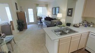 This single-story space showcases an open-concept design that seamlessly connects the kitchen and living area, creating a cohesive and functional layout. The kitchen features a peninsula with a double-basin stainless steel sink and light-toned cabinetry, providing ample storage and counter space while maintaining a clear sightline into the living area. Neutral countertops complement the warm, understated color palette found throughout the space. The living area is carpeted in a plush, light-toned textile that adds warmth underfoot. Large windows and a glass-paneled door flood the space with natural light, offering views of the outdoors and enhancing the open, airy ambiance. Sheer green-accented curtains frame the windows, adding a subtle touch of color without disrupting the room's soft, neutral tone. Framed artwork on the walls contributes to the finished, well-appointed feel of the space. The overall atmosphere is bright and welcoming, with a layout well-suited for both everyday living and casual entertaining. The open flow between the kitchen and living room ensures easy interaction between spaces, making the home feel connected and spacious.