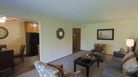 This single-floor space offers a straightforward, functional layout with clearly defined living and kitchen areas. The living room is carpeted and furnished with patterned upholstered armchairs arranged around a dark rectangular coffee table, creating a comfortable seating arrangement. A decorative circular mirror adorns one of the light-colored walls, while a framed piece of artwork adds visual interest on an adjacent wall. The neutral wall tones throughout contribute to a clean, understated aesthetic.

A doorway visible from the living area leads to an additional room, suggesting a connected layout that allows for easy movement between spaces. The kitchen, partially visible from the living room, features dark cabinetry and a black refrigerator, contrasting with the lighter tones of the surrounding walls. The transition between the carpeted living space and the hard flooring of the kitchen area provides a natural visual boundary between the two zones.

Overall, the space presents a modest, practical interior with standard finishes, offering a livable environment with a simple, cohesive design throughout its single level.