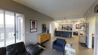 This single-floor, 2-bedroom, 2-bathroom residence features an open-concept design that seamlessly connects the living area to the kitchen, creating a spacious and functional environment. The living room is anchored by a sleek black leather sofa and a navy blue accent chair, complemented by light hardwood-style flooring that runs throughout the main living spaces. Gray-painted walls provide a modern, neutral backdrop, enhanced by curated wall art that adds personality to the space.

The kitchen is a standout feature, showcasing dark cabinetry with stainless steel appliances, including a refrigerator and range. A dark-topped island with bar stools serves as both a prep area and casual dining spot, making it ideal for entertaining. Recessed lighting throughout the kitchen ensures ample illumination.

A door visible from the living area suggests convenient access to a patio or balcony, allowing for natural light to filter into the space. The overall ambiance is contemporary and cohesive, with a thoughtful blend of functional furnishings and modern design elements, making this residence well-suited for comfortable, everyday living.