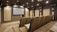 This private home theater is thoughtfully designed to replicate the full cinematic experience within a dedicated, single-level screening room. The space is anchored by a large projection screen positioned at the front wall, flanked by acoustic panels finished in neutral tones and framed with dark wood trim — a detail that repeats throughout the room to create a cohesive, upscale aesthetic.

Rows of plush, tan leather stadium-style seats are arranged on a tiered floor plan, ensuring unobstructed sightlines from every position. Each seat is equipped with built-in cup holders and padded armrests, prioritizing comfort during extended viewing sessions. A ceiling-mounted projector serves as the room's central technical component, complemented by a media equipment stack positioned beneath the screen.

The walls are lined with decorative acoustic panels framed by dark pilasters, which both enhance sound quality and contribute to the theater's polished interior design. Recessed spotlights are embedded in the dark-painted ceiling, providing subtle, controllable ambient lighting that reinforces the immersive atmosphere. The floor is covered in a patterned carpet with a geometric motif, adding texture and warmth to the space. Together, these elements create a sophisticated, purpose-built environment ideal for film screenings, presentations, or private entertainment.