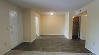 This single-story space presents a clean and functional layout with a neutral, move-in-ready aesthetic. The main living area features dark wood-look flooring that contrasts with the warm beige walls and white baseboards, creating a grounded, cohesive look throughout. The textured ceiling adds subtle visual depth, while a flush-mount ceiling light provides central illumination to the space.

Two distinct openings branch off from the main area. To the left, a doorway leads to what appears to be a bathroom, glimpsed by white cabinetry and light-colored tile or flooring. To the right, an open entryway reveals a spacious walk-in closet outfitted with built-in shelving, offering generous and well-organized storage potential.

The overall ambiance is understated and practical, with a simple color palette that lends itself well to a variety of furnishing styles. The absence of clutter and the open flow of the layout make the space feel airy and adaptable. The combination of hardwood-style flooring and ample storage options makes this a versatile and functional living space.
