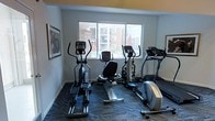 This fitness center is a well-appointed, single-floor workout space designed to meet essential cardio training needs. The room features a selection of cardio equipment, including elliptical trainers, a recumbent stationary bike, and a treadmill, arranged to maximize the available floor space while maintaining comfortable circulation between machines. The flooring consists of a durable, patterned carpet in muted blue and gray tones, providing both cushioning and a clean aesthetic underfoot. A large, multi-pane window along one wall allows ample natural light to fill the room and offers views of the surrounding building exterior and outdoor areas, creating an open and airy atmosphere during workouts. The walls are painted in a neutral light gray, accented by a warm yellow-toned upper border, contributing to an energizing yet approachable environment. Framed artwork on the walls adds a touch of character to the space. The overall ambiance is clean, functional, and welcoming, making it a convenient and practical facility for residents or guests seeking a dedicated space for daily cardiovascular exercise.