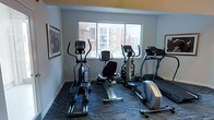 This fitness center is a well-appointed, single-floor workout space designed to meet essential cardio training needs. The room features a selection of cardio equipment, including elliptical trainers, a recumbent stationary bike, and a treadmill, arranged to maximize the available floor space while maintaining comfortable circulation between machines. The flooring consists of a durable, patterned carpet in muted blue and gray tones, providing both cushioning and a clean aesthetic underfoot. A large, multi-pane window along one wall allows ample natural light to fill the room and offers views of the surrounding building exterior and outdoor areas, creating an open and airy atmosphere during workouts. The walls are painted in a neutral light gray, accented by a warm yellow-toned upper border, contributing to an energizing yet approachable environment. Framed artwork on the walls adds a touch of character to the space. The overall ambiance is clean, functional, and welcoming, making it a convenient and practical facility for residents or guests seeking a dedicated space for daily cardiovascular exercise.