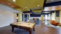 Located at 2200 Bellamy Ct in Greenville, NC, this single-floor clubhouse and amenity space is designed with an energetic, social atmosphere that encourages community engagement. The space is anchored by a bold, contemporary aesthetic, featuring a striking color palette of deep navy blue, olive green, and warm neutral tones across the walls, ceilings, and architectural accents.

The centerpiece of the main lounge area is a full-size billiards table, positioned beneath a multi-tiered ceiling with recessed lighting that creates a dynamic visual overhead. The "#BELLAMY" branding emblazoned on the navy accent wall adds a modern, social-media-inspired touch that reflects the community's identity. Adjacent to the pool table is a comfortable lounge zone furnished with plush sofas and armchairs, ideal for casual conversation and relaxation.

The space flows seamlessly into additional areas, including a dining or café-style seating section visible in the background, offering versatile options for residents. A kitchenette or refreshment area is also accessible, adding functional convenience to the space. Large windows allow natural light to filter in, enhancing the open, welcoming ambiance. Overall, this amenity space is thoughtfully designed to serve as a lively social hub for its residents.