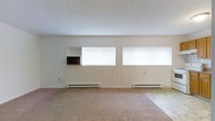 This single-floor space presents a straightforward, open layout centered around a spacious main room finished with wall-to-wall carpeting in a neutral beige tone. The room is illuminated by natural light streaming through multiple horizontal windows fitted with white blinds, positioned along one wall to create a bright, airy atmosphere. A textured popcorn ceiling adds a classic touch overhead, while the smooth, neutral-toned walls provide a clean backdrop throughout.

A built-in wall niche with a small open shelf adds a subtle architectural detail, offering a functional display or storage element without occupying floor space. Baseboard heating units run along the base of the exterior wall beneath the windows, providing an efficient heating solution distributed evenly across the room. The overall color palette is understated and neutral, with beige carpeting and off-white walls contributing to a calm, versatile environment. The space appears well-suited for a variety of uses, with its open floor plan allowing for flexible furniture arrangements. The combination of natural light, baseboard heat, and built-in shelving makes this a practical and functional living area.