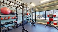 Located at 3815 Maypop Cir in Myrtle Beach, SC, this fitness center is a compact yet thoughtfully designed exercise space that maximizes every square foot for a comprehensive workout experience. The room features soft sage-green walls, recessed lighting, and a tray ceiling detail that adds architectural character and a sense of height to the space.

Large windows line one wall, flooding the room with natural light and offering views of the surrounding outdoor area, including a pool and landscaping. Full-length mirrors on an adjacent wall visually expand the space and allow users to monitor their form during exercise.

The fitness area is divided into two functional zones. A cardio section is equipped with multiple TRUE brand treadmills and additional cardio machines, including a rowing station and a cable machine with adjustable pads. The strength and functional training zone features a robust multi-use power rack system branded with Throwdown, complete with integrated storage shelving holding medicine balls, stability balls, kettlebells, dumbbells, resistance bands, and weight plates. A flat/adjustable weight bench and barbell complete the free weights setup.

Durable rubberized flooring covers the entire exercise area, providing safety and cushioning. A glass sliding door provides access to an adjoining room, contributing to the open, accessible feel of this well-appointed fitness facility.
