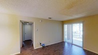 This single-floor residence offers a clean and functional layout designed for comfortable everyday living. The main living area features rich, dark hardwood flooring that extends throughout, providing a warm and grounded aesthetic that contrasts neatly with the light-toned walls painted in a soft, neutral yellow-beige hue. A textured popcorn ceiling adds a classic architectural detail overhead, while a central HVAC vent ensures year-round climate comfort. Natural light floods the living space through a full-height sliding glass door fitted with vertical blinds, offering a direct view and access to an outdoor area — ideal for relaxing or entertaining. The open wall space provides flexibility for furniture arrangement, making the living area highly adaptable. A connecting hallway leads to the remaining rooms of the unit, with a lighted ceiling fixture illuminating the passage. The hallway access point hints at a well-organized floor plan with clearly defined zones for living, sleeping, and utility. Overall, the space strikes a balance between simplicity and practicality, with durable finishes and a layout that maximizes usability within a compact, single-level footprint.