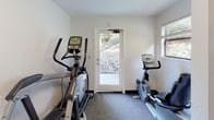 This fitness center is a compact, single-floor exercise space designed to support a well-rounded workout routine. The room features neutral white walls, dark baseboards, and carpet flooring throughout, creating a clean and functional environment. A large, dark-framed full-length mirror is mounted on one wall, enhancing the sense of space while allowing users to monitor their form during exercise.

The centerpiece of the strength training area is a multi-functional cable machine, equipped with a variety of attachments including a rope pull-down and adjustable cable arms, suitable for a wide range of resistance exercises. An adjustable weight bench is positioned nearby for additional strength work. A small wall-mounted shelf and a waste bin are also present to keep the space organized.

The cardio area includes an elliptical trainer and a recumbent stationary bike, both positioned near a glass exterior door and a window fitted with blinds, allowing natural light to filter in and offering views of an outdoor rocky landscape. A seating chair is available within the space.

Motivational wall art displaying the phrase "Strive for Progress, Not Perfection" adds an encouraging ambiance to this practical and accessible fitness facility.