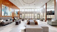 Located at 180 Woodruff Rd in Greenville, SC, this single-floor living space offers a refined and modern interior defined by its striking architectural details and thoughtfully curated design. The centerpiece of the room is a floor-to-ceiling warm wood accent wall, which serves as a bold backdrop for large-scale abstract artwork in rich tones of teal, gold, and navy — immediately drawing the eye and setting an sophisticated tone throughout.

The space features soaring ceilings that amplify the sense of openness, complemented by large multi-pane windows dressed in sheer, light-filtering curtains that flood the interior with abundant natural light and provide views of the surrounding greenery. The large-format tile flooring, laid in a subtle checkerboard pattern, adds visual interest while maintaining a clean, contemporary aesthetic.

Seating areas are arranged to encourage conversation and comfort, with plush gray sofas and accent chairs positioned around glass-topped coffee tables with chrome frames. A wall-mounted flat-screen television is framed by a mirrored feature wall, adding depth to the room. Built-in shelving with curated décor accents and a pair of statement entry doors with geometric metallic detailing further enhance the space's polished, upscale character. Lush indoor plants add a welcoming, organic touch throughout.