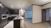 This single-floor space showcases a thoughtfully designed open-concept layout that seamlessly connects the kitchen and living areas. The kitchen is a standout feature, equipped with rich dark espresso cabinetry, a classic tile backsplash, and a granite-topped island with an undermount sink that serves as both a functional workspace and a natural gathering point. Pendant lighting hangs elegantly above the island, complementing the recessed track lighting overhead. Light-toned hardwood flooring extends throughout the space, creating a bright and cohesive aesthetic that contrasts beautifully with the darker cabinetry. The open layout allows for a fluid transition between the kitchen and the adjacent living area, which benefits from the spacious feel created by the clean lines and neutral wall tones. A hallway visible from the main living space leads to additional rooms, including what appears to be a bathroom, maintaining the same modern design language found throughout. Crown molding details and accent trim add refined architectural touches to the overall interior. The result is a well-appointed, contemporary space that balances style and functionality in a clean, modern presentation.