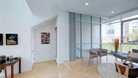 This three-story space showcases a contemporary design aesthetic defined by clean lines, bright white walls, and light hardwood flooring throughout its common areas. Large floor-to-ceiling glass partitions with industrial-style steel framing flood the interior with natural light, creating an open, airy atmosphere. Pleated sheer curtains offer flexible privacy control along the expansive glazed walls, while recessed lighting ensures a well-lit environment throughout.

A seating area adjacent to the glass wall provides a comfortable lounge space, complemented by a hallway featuring angled ceiling details that add architectural interest. A bedroom features plush carpeting, a large window that draws in abundant natural light, and views of the surrounding urban landscape, contributing to a bright and restful retreat.

One of the standout features of this space is the private rooftop terrace on the uppermost level, offering sweeping views of the surrounding cityscape under open skies — ideal for outdoor relaxation. The ground level includes a spacious attached garage with an automatic door opener, providing secure, convenient parking with direct interior access. Together, these elements create a well-appointed, modern urban residence.