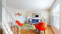 This two-floor space offers a well-organized layout with distinct areas across each level. The main living area is a standout feature, showcasing warm hardwood flooring, large windows fitted with blinds that invite generous natural light, and a cheerful, contemporary aesthetic. Bold orange accent chairs, a blue sofa, and a vivid gray-and-yellow area rug centered beneath a glass coffee table create an energetic yet cohesive atmosphere. A framed mirror adorns the wall, amplifying the sense of space. A staircase with white railings connects the two floors.

The kitchen is outfitted with white cabinetry, light granite-style countertops, and standard appliances including a range and refrigerator, providing ample storage and a clean, practical workspace.

The bedrooms are carpeted for comfort. One bedroom features a decorative lattice-panel room divider and soft, neutral tones, while another is appointed with classic wrought-iron furnishings and warm wood accents. The bathrooms include a full bath with a shower enclosure and a secondary half bath featuring a textured accent wall. A dedicated laundry room houses a full-sized washer and dryer, adding everyday convenience to this well-appointed space.
