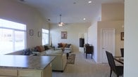 This single-story residence showcases an open-concept layout that connects the kitchen and living areas into one cohesive, functional space. The living room is defined by impressively tall ceilings that enhance the sense of spaciousness and airiness throughout. A ceiling fan is centrally mounted, contributing both to air circulation and to the room's understated aesthetic. Recessed lighting complements the natural light entering through windows along the far wall.

The living area is finished with plush wall-to-wall carpeting and features a sectional sofa arrangement centered around a glass-top coffee table, anchored by a decorative area rug. Warm accent pillows in earth tones add depth to the neutral color palette that runs throughout the space. A dark wood dresser and side cabinet add contrast against the light beige walls.

The kitchen, partially visible in the foreground, includes granite countertops and light wood cabinetry. A hallway visible in the background suggests additional rooms beyond the main living space. A white paneled door leads to an exterior exit. The overall ambiance is calm, neutral, and well-proportioned, offering a comfortable and inviting environment.