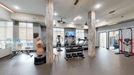 Located at 2301 E Tenth St in Greenville, NC, this fitness center offers a thoughtfully designed, two-zone workout facility spanning a generously sized single floor. The main exercise area is distinguished by decorative stone-clad columns, speckled rubberized flooring, and large windows that flood the space with natural light. Ceiling fans provide comfortable airflow throughout, while recessed lighting and pendant fixtures ensure bright, even illumination. The main zone is comprehensively outfitted with a full dumbbell rack, a multi-station cable machine, a red power rack with a weight bench, medicine ball storage, stability balls, treadmills, ellipticals, a stair climber, and a wall-mounted flat-screen TV for entertainment during workouts. A striking large-scale mural adds an artistic focal point to one wall, enhancing the space's modern aesthetic. The secondary room features a striking horizontal wood-slat accent wall, soft blue rubberized flooring, a split-system climate control unit, and equipment including a rowing machine and spin bike, making it ideal for focused cardio or recovery sessions. A glass-paneled door connects this area to the outdoors, reinforcing the bright, open atmosphere of the overall facility.