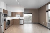 This contemporary single-floor residence showcases a clean, modern design throughout. The kitchen is a standout feature, equipped with stainless steel appliances including a refrigerator, electric range, microwave, and dishwasher. Rich wood-toned cabinetry with frosted glass upper cabinets frames the space, complemented by granite countertops and a white tile backsplash. Large-format tile flooring adds a polished, cohesive feel.

The open living area is anchored by a striking slate-tiled fireplace and benefits from tall ceilings with track lighting that creates an airy, well-lit atmosphere. Floor-to-ceiling wood-framed glass doors lead to an outdoor area, inviting natural light and views of the surrounding greenery.

The bedrooms feature soft carpeting and large windows that frame outdoor views, providing a calm and comfortable retreat. Each room is finished with clean white walls and dark baseboards that lend a crisp, contemporary edge.

Two bathrooms serve the residence. One includes a soaking tub with a dramatic dark tile surround accented by wood-toned horizontal banding. The second features a walk-in glass shower, a wood-cabinet vanity with a granite countertop, and a large mirror. Both bathrooms are finished with coordinating tile flooring for a cohesive, refined aesthetic throughout.