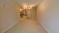 This single-floor space features an open-concept design that seamlessly connects the living area to the kitchen and entryway. The living area is finished with neutral-toned wall-to-wall carpeting, creating a comfortable and cohesive feel throughout. Clean white baseboards frame the space, contributing to a tidy, well-maintained aesthetic.

The kitchen is positioned toward the rear of the unit and is outfitted with warm honey-toned wood cabinetry, dark countertops, and stainless steel appliances, including a microwave and stove. The kitchen layout provides functional counter and storage space, making it practical for everyday use.

A white entry door with a single sidelight panel anchors the entryway, which is finished with tile flooring that transitions into the carpeted living space. Overhead, track-style and flush-mount light fixtures provide consistent illumination throughout the common areas. The neutral beige wall tones throughout lend a warm, inviting atmosphere, and the open sightlines between rooms contribute to an airy, spacious feel despite the compact single-floor footprint.