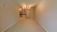 This single-floor space features an open-concept design that seamlessly connects the living area to the kitchen and entryway. The living area is finished with neutral-toned wall-to-wall carpeting, creating a comfortable and cohesive feel throughout. Clean white baseboards frame the space, contributing to a tidy, well-maintained aesthetic.

The kitchen is positioned toward the rear of the unit and is outfitted with warm honey-toned wood cabinetry, dark countertops, and stainless steel appliances, including a microwave and stove. The kitchen layout provides functional counter and storage space, making it practical for everyday use.

A white entry door with a single sidelight panel anchors the entryway, which is finished with tile flooring that transitions into the carpeted living space. Overhead, track-style and flush-mount light fixtures provide consistent illumination throughout the common areas. The neutral beige wall tones throughout lend a warm, inviting atmosphere, and the open sightlines between rooms contribute to an airy, spacious feel despite the compact single-floor footprint.