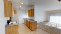 This single-floor residence offers a practical and welcoming layout centered around an open-concept kitchen and living area. The kitchen is equipped with warm honey-toned wood cabinetry spanning both upper and lower sections, providing generous storage space. A double-basin sink is positioned along the main counter run, complemented by dark granite-style countertops that offer a subtle contrast to the light-colored walls. White appliances, including a refrigerator, electric range, and dishwasher, complete the functional kitchen setup. Light tile flooring runs throughout the kitchen area, creating a clean and easy-to-maintain surface underfoot. The kitchen flows seamlessly into an adjoining living area, which features plush carpeting and benefits from natural light streaming through a window. Recessed lighting in the kitchen adds a modern touch and ensures the space is well-illuminated. The crisp white walls throughout lend a bright, neutral backdrop that makes the space feel open and airy. The overall layout and design reflect a straightforward, functional aesthetic that is both comfortable and easy to personalize.