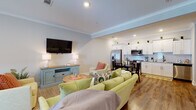This single-floor space offers a well-organized, open-concept layout that seamlessly connects the living, dining, and kitchen areas, creating an inviting and functional environment. Throughout the space, warm hardwood-style flooring and crown molding provide a cohesive, polished aesthetic.

The living area is generously proportioned, featuring recessed lighting and a wall-mounted flat-screen television above a coastal-style blue media console. Comfortable seating, including a green sofa and accent chairs with coral pillows, establishes a relaxed, welcoming atmosphere. A glass-paned door allows natural light to filter into the space.

The kitchen is well-appointed with white shaker-style cabinetry, a classic subway tile backsplash, stainless steel appliances including a French door refrigerator, range, microwave, and dishwasher, and ample counter space. A dedicated dining area with a dark table and chairs sits adjacent to the kitchen.

The primary bedroom is spacious, featuring multiple windows, recessed lighting, crown molding, and an en-suite bathroom. The bathroom includes a tiled walk-in shower, a white vanity, and hardwood-style flooring. An additional, cozier bedroom provides comfortable accommodations with a window and closet access.