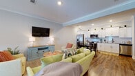This single-floor space offers a well-organized, open-concept layout that seamlessly connects the living, dining, and kitchen areas, creating an inviting and functional environment. Throughout the space, warm hardwood-style flooring and crown molding provide a cohesive, polished aesthetic.

The living area is generously proportioned, featuring recessed lighting and a wall-mounted flat-screen television above a coastal-style blue media console. Comfortable seating, including a green sofa and accent chairs with coral pillows, establishes a relaxed, welcoming atmosphere. A glass-paned door allows natural light to filter into the space.

The kitchen is well-appointed with white shaker-style cabinetry, a classic subway tile backsplash, stainless steel appliances including a French door refrigerator, range, microwave, and dishwasher, and ample counter space. A dedicated dining area with a dark table and chairs sits adjacent to the kitchen.

The primary bedroom is spacious, featuring multiple windows, recessed lighting, crown molding, and an en-suite bathroom. The bathroom includes a tiled walk-in shower, a white vanity, and hardwood-style flooring. An additional, cozier bedroom provides comfortable accommodations with a window and closet access.