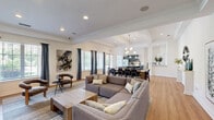 Located in Ooltewah, Tennessee, this single-floor space offers an open-concept layout that seamlessly connects the living room, dining area, and kitchen. Light hardwood flooring flows throughout, complemented by crisp white walls and elegant crown molding that lend a clean, modern aesthetic.

The living area is bright and welcoming, featuring a tray ceiling with recessed lighting, large windows dressed with charcoal drapes that invite natural light, and ample room for comfortable seating arrangements. A wall-mounted television and a warm color palette create a relaxed yet refined atmosphere.

The dining area stands out with its dramatic exposed ceiling beams and a striking contemporary chandelier featuring gold-toned arms and globe lights. A generous dining table comfortably seats multiple guests, making this space well-suited for gatherings.

The kitchen is thoughtfully designed with dark grey cabinetry, stainless steel appliances, and a large island topped with richly veined granite countertops. Pendant lighting above the island adds a modern touch, while abundant natural light streams in through wide windows. A sleek, floor-to-ceiling fireplace framed in neutral tile serves as an architectural focal point between the kitchen and adjoining areas, adding both warmth and visual interest to the overall space.
