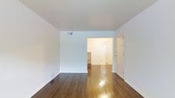This single-floor space presents a clean, minimalist aesthetic defined by its bright white walls and ceilings that create an airy, open atmosphere throughout. The main living area is immediately notable for its rich, polished hardwood floors that run continuously through the space, reflecting light and adding warmth to the otherwise neutral palette. The floors display a classic wood grain with a glossy finish, lending the space a timeless character.

A centrally positioned interior opening connects the main living area to an adjoining space, providing a sense of depth and visual flow through the unit. Beyond this opening, a well-lit entryway is visible, featuring a windowed door that allows natural light to filter inward, brightening the interior. The entryway also includes what appears to be a coat closet or storage area, offering practical functionality near the entrance.

Ceiling-mounted HVAC vents are integrated cleanly into the white ceiling, maintaining the uncluttered look of the space. Baseboard trim runs consistently along the lower walls, complementing the hardwood flooring. The overall ambiance is fresh, neutral, and move-in ready, providing a versatile canvas well-suited for a variety of furnishing styles.