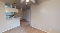 This single-floor space offers a clean, functional layout designed for comfortable everyday living. The main living area features warm-toned ceramic tile flooring throughout, providing a durable and easy-to-maintain surface. A ceiling fan with a light fixture is centrally mounted, ensuring consistent airflow and ambient lighting across the space.

A partial kitchen bar counter is visible along one side, creating a natural division between the living area and the kitchen space. The counter features a light-colored surface supported by a white base, offering a convenient area for casual dining or meal preparation. A pendant light fixture is also present near the kitchen area, adding focused task lighting.

The walls are finished in a neutral beige tone, contributing to a bright and welcoming atmosphere. White baseboards provide a clean contrast along the perimeter of the room. A hallway visible in the background leads to additional areas of the space, with a thermostat panel mounted on the wall nearby. Electrical outlets are conveniently positioned throughout, and a fire extinguisher is noted near the kitchen area, reflecting attention to safety. Overall, the space presents a straightforward, well-maintained interior with practical finishes suited for a variety of occupants.