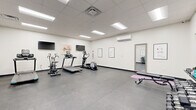 Located at 501 State St in Schenectady, New York, this fitness center is a thoughtfully designed, single-room exercise facility that provides a complete workout environment. The space features a drop-panel ceiling with recessed fluorescent lighting that ensures bright, even illumination throughout, while a wall-mounted split air conditioning unit maintains a comfortable climate year-round.

The layout is divided into distinct functional zones. The cardio area is well-stocked with multiple treadmills, an elliptical trainer, a stationary air bike, and a cycling bike, each accompanied by wall-mounted flat-screen televisions for an engaging workout experience. The strength training area houses a three-tier dumbbell rack with a full range of free weights, kettlebells, and an adjustable workout bench, providing ample options for resistance training. Framed exercise instructional diagrams are displayed along the white walls, serving as a practical reference for users.

The durable dark rubber-tile flooring covers the entire space, offering stability and impact absorption suited for all types of exercise. A full-length mirror is mounted on one wall, allowing users to monitor their form. The facility is accessible via a dedicated exterior entrance and connects to an adjacent room visible through an interior door. The clean, minimal aesthetic and efficient use of space make this fitness center a fully functional and inviting place to train.