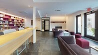 Located at 3200 Washington St in Boston's Jamaica Plain neighborhood, this single-floor community amenity space combines contemporary design with functional comfort. The open layout features large-format dark slate tile flooring throughout, creating a sleek, cohesive foundation that anchors the entire space.

The main lounge area is anchored by a modern gas fireplace set into a clean white brick surround with a built-in display niche above, providing a warm and inviting focal point. Plush gray and deep purple sofas are arranged to encourage conversation, complemented by vibrant mustard yellow accent chairs and jewel-toned ottomans in burgundy and purple that inject pops of color throughout.

A well-equipped kitchenette area features light blue-gray cabinetry, open wood shelving stocked with dishware, a microwave, and ample counter space, all set against a decorative patterned backsplash. Adjacent to this is a dining and media area with a large projection screen, making it well-suited for group gatherings or presentations.

Natural light flows generously through floor-to-ceiling black-framed glass doors and windows, which open directly to an outdoor patio furnished with seating. Framed black-and-white artwork adorns the crisp white walls, while recessed lighting and track fixtures provide flexible illumination. A built-in shelving unit adds a functional library element to round out this versatile communal space.