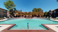 This community amenity space combines an expansive outdoor pool area with a beautifully designed interior clubhouse. The outdoor pool is generously sized, featuring a classic brick surround deck with metal handrails and decorative tiled accents along its curved walls. Surrounding the pool are lounge chairs and mature trees that provide natural shade and a vibrant backdrop, with the residential buildings visible in the distance beyond a wrought iron fence.

The interior clubhouse is equally impressive, boasting soaring vaulted ceilings that create an open, airy atmosphere throughout. Light-toned tile flooring runs continuously through the space, enhancing the bright, clean aesthetic. Recessed lighting and natural light from large windows illuminate the interior. A welcoming lounge area features plush, neutral-toned sectional sofas accented with patterned throw pillows, arranged around a sleek coffee table atop a decorative area rug. Adjacent to the lounge, a dining area with upholstered chairs surrounds a round table near the entrance. Classic furnishings, including an ornate dark wood console and a traditional-style accent chest, add refined character throughout. The entry foyer provides a gracious arrival experience, anchored by an elegantly detailed dark wood dresser beneath a gridded window.