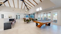 Located in Wildwood, Florida, this generously sized single-floor clubroom offers an impressive and versatile gathering space designed for both leisure and socializing. The architectural centerpiece is the dramatic vaulted ceiling, accented by exposed natural wood beams and striking geometric chandelier pendants that cast warm ambient light throughout the open interior. Large-format light tile flooring spans the entire space, enhancing its bright and airy atmosphere.

The layout is thoughtfully divided into distinct activity zones. A full-size pool table anchors one side of the room, while a comfortable lounge area features a plush sectional sofa paired with a wood-accent media wall fitted with a large flat-screen TV. A set of green velvet dining chairs and sleek tables create an inviting gathering area in the center, complemented by a high-top bistro table with leather-style bar stools positioned near floor-to-ceiling windows. A long bar-height counter along one wall provides additional seating.

Abundant natural light pours in through large windows and glass French doors that open to an outdoor area, offering views of the surrounding greenery. The overall design balances modern finishes with warm, natural materials, creating a welcoming and stylish environment suited for community events and everyday relaxation.