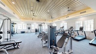Located on Mulberry Blvd in Port Wentworth, GA, this well-appointed fitness center offers a generously sized, single-floor workout facility designed to support a comprehensive exercise experience. The space is defined by its standout architectural feature: a warm, wood-planked tray ceiling accented with indirect LED lighting and multiple ceiling fans, creating an inviting yet energizing atmosphere. Crisp white walls and large windows lining multiple sides of the room flood the interior with natural light, enhancing the open, airy feel of the space.

The fitness floor is equipped with an extensive selection of commercial-grade machines and free weights. Cardio equipment includes a row of treadmills and additional aerobic machines, while the strength training area provides cable machines, a multi-functional rack system, selectorized weight stations, a rowing machine, and a full dumbbell rack with benches for free weight exercises. The layout is thoughtfully organized to allow clear zones for different training modalities, ensuring ample circulation space between equipment.

Durable, low-pile carpet flooring covers the workout area, offering comfort underfoot while maintaining a clean, modern aesthetic. A wall-mounted flat-screen TV and posted facility guidelines round out the practical amenities. The overall ambiance is bright, clean, and professional, reflecting a commitment to providing a high-quality fitness environment.