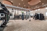 This expansive fitness center in Idaho Falls, Idaho, offers a comprehensive, single-floor layout designed to support a wide range of athletic and wellness activities. The main gym area features soaring ceilings with warm terracotta-toned accent soffits, recessed lighting, and large ceiling fans that contribute to a modern, energetic atmosphere. Clerestory windows line the upper walls throughout, inviting abundant natural light while maintaining privacy. The strength training zone is well-appointed with a full rack of dumbbells, multiple cable machines, weight benches, and a dedicated functional training rig equipped with battle ropes, a heavy bag, gymnastic rings, and suspension trainers. A rubber flooring section anchors the free weights area, while plush carpet tile extends through the rest of the gym. The cardio area houses a row of treadmills, ellipticals, stair climbers, and stationary bikes, all positioned near windows for natural light during workouts. Large mirrors amplify the sense of space throughout the weight room. Adjacent to the gym is a full-size indoor multi-sport court with a regulation basketball hoop, badminton net, and colorful geometric wall murals, illuminated by bright overhead lighting and high clerestory windows. Padded wall panels and a hardwood-bordered sport floor complete this versatile court space.