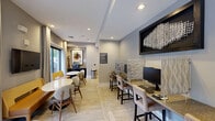 Located at 131 Poindexter Drive in Charlotte, NC, this well-appointed single-floor amenity space offers a thoughtfully designed environment that balances functionality with contemporary style. The main lounge and dining area features light wood-look flooring, recessed lighting, and large floor-to-ceiling windows dressed with floor-length drapery, flooding the space with natural light and providing views of the tree-lined streetscape outside. Marble-top round tables paired with mustard yellow upholstered benches and mid-century modern chairs create an inviting and stylish atmosphere. Wall-mounted televisions and curated abstract artwork add visual interest throughout. A dedicated business center is seamlessly integrated into the open layout, offering counter-height workstations with desktop computers, granite surfaces, and comfortable seating — ideal for productivity. A private conference room anchors one end of the space, distinguished by a bold geometric-patterned accent wall in warm earth tones, a long light-wood conference table, a mounted flat-screen display, an elegant multi-arm chandelier, and a private phone booth for focused calls. Neutral gray walls and patterned carpet complete the room's polished, professional look. Together, these areas form a versatile and refined communal space.