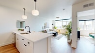 This single-floor residence showcases a thoughtfully designed open-concept layout that seamlessly connects the kitchen and living areas. The kitchen is outfitted with white cabinetry featuring matte black hardware, a crisp white countertop island with seating for three, stainless steel appliances including a full-size refrigerator and built-in microwave, and a black-finish faucet over an undermount sink. Pendant lighting with soft globe shades adds warmth overhead, while warm-toned hardwood flooring runs throughout the space.

The living area is bathed in natural light through large windows that frame elevated city views of the surrounding urban skyline. A curated gallery wall and comfortable seating arrangements create an inviting atmosphere. An additional flexible room functions as a home office or secondary sitting space and benefits from the same generous window views.

The bedroom is a calm retreat, featuring wide windows that capture sweeping city vistas and allow abundant daylight. Clean white walls enhance the airy feel of the room.

The bathroom features a glass-enclosed walk-in shower lined with vertically stacked white subway tiles and chrome fixtures, delivering a clean, modern aesthetic consistent throughout the residence.